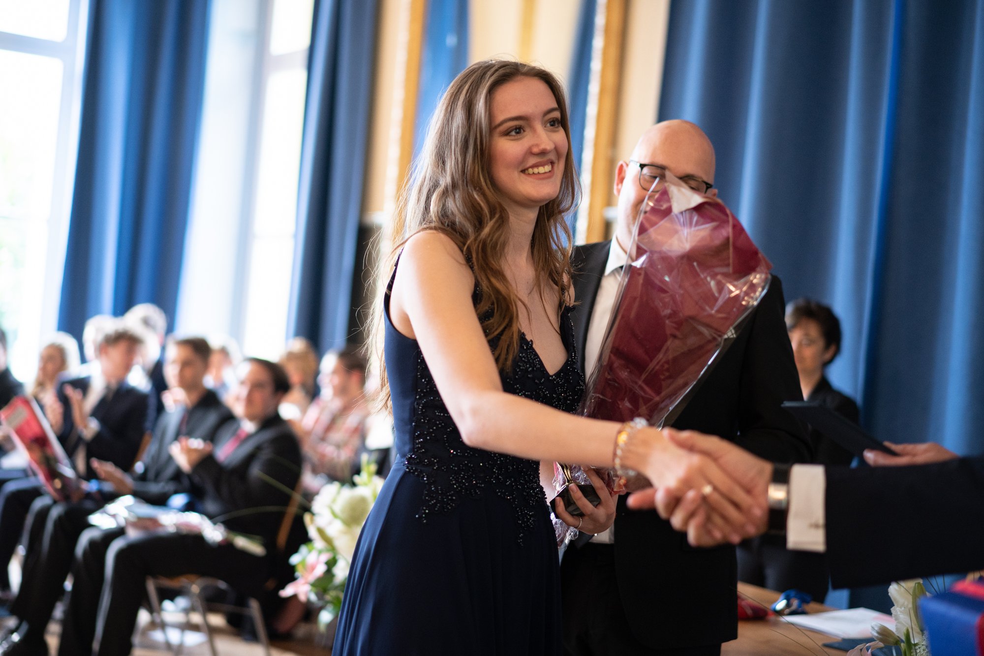 A female student shakes hand on a graduation day. Other students sit in the background.