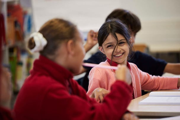 Zwei Grundschülerinnen, die sich in einer Klasse mit einem Grundschüler im Hintergrund unterhalten und Spaß haben
