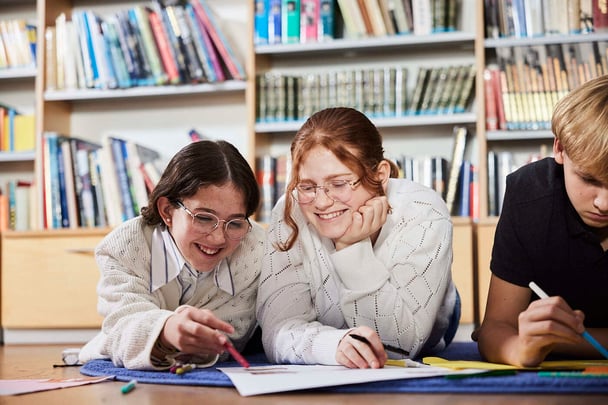 Zwei Schülerinnen und ein Schüler arbeiten auf dem Boden einer Bibliothek an etwas.
