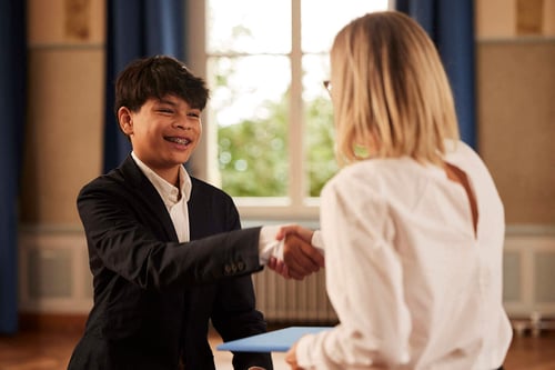 A boy student shakes hand with a female teacher in an aula.