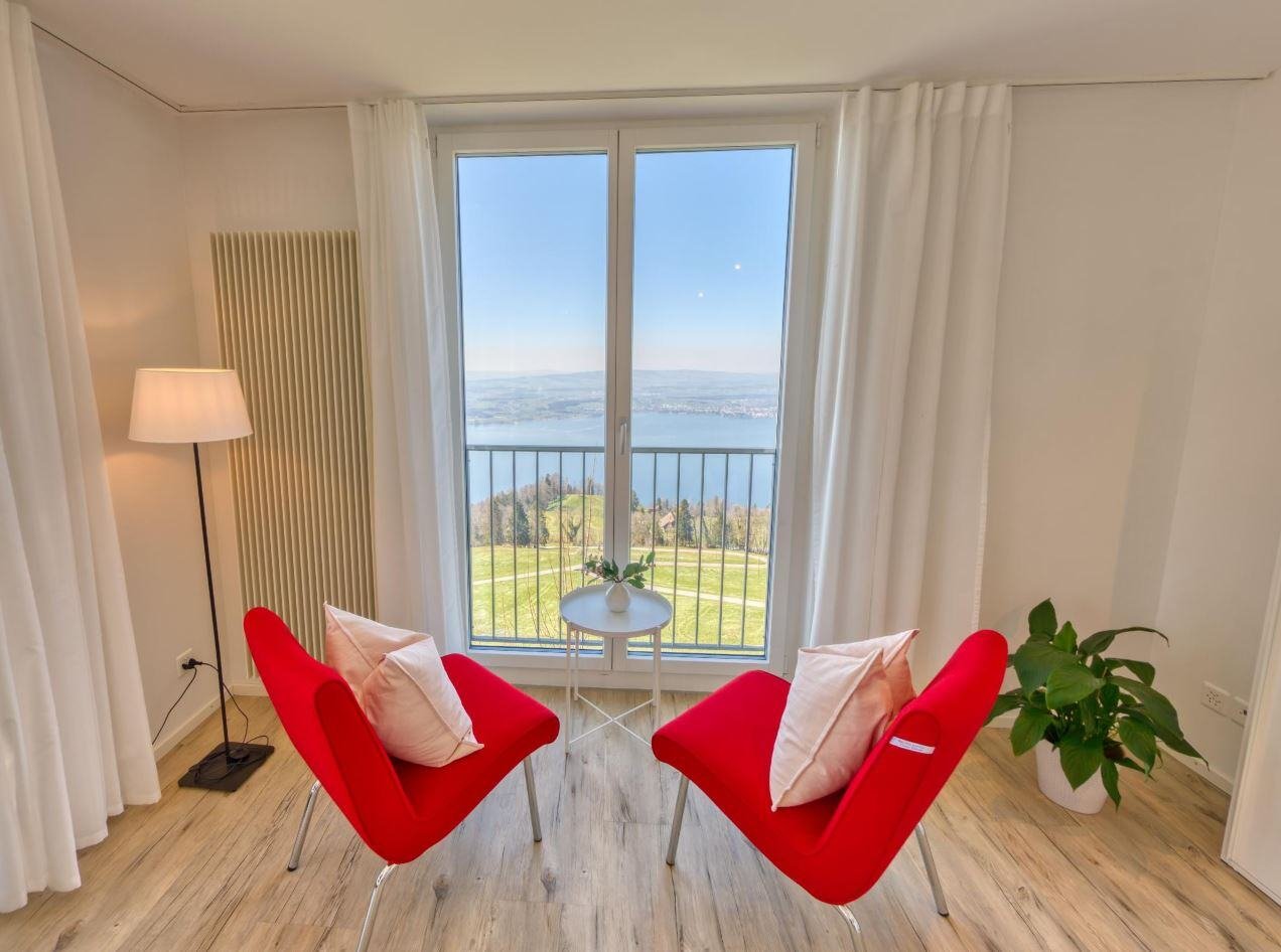 Ein offenes Fenster eines hellen, luftigen Zimmers mit Blick auf den Zugersee, zwei rote Sofas mit einem kleinen Tisch vor dem Fenster.