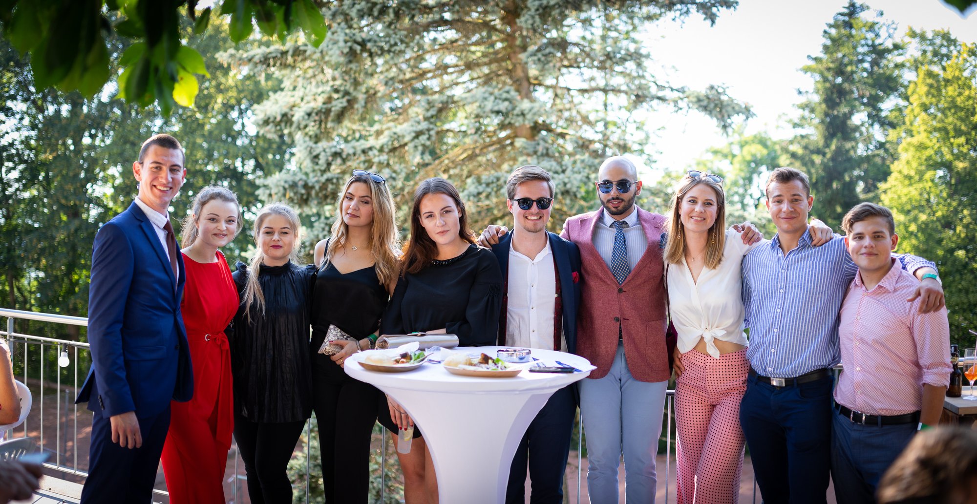 Eine Gruppe von Studenten in formellen Outfits posiert glücklich vor der Kamera in einer Außenumgebung.