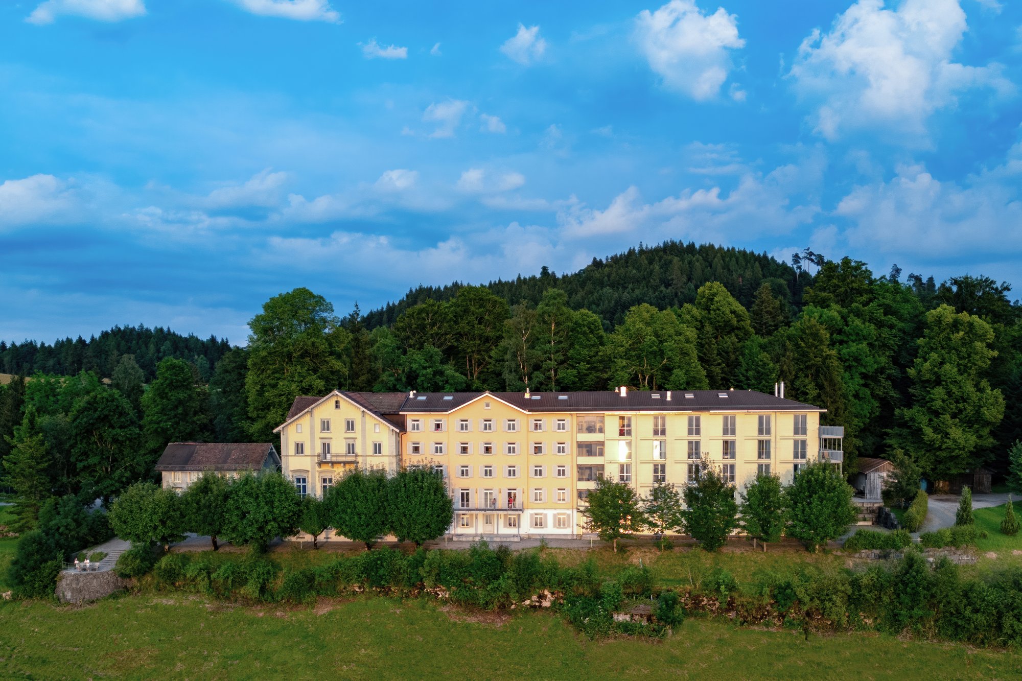 Ein hellgelb gestrichenes langes Gebäude, umgeben von Wald und Bäumen.