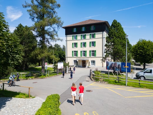 Ein cremefarbenes Gebäude mit grünen Fenstern und braunem Dach auf einem üppig grünen Campus, auf dem einige Studenten herumlaufen.