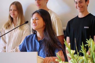 A girl student speaks in front of a microphone with a group of other student behind her