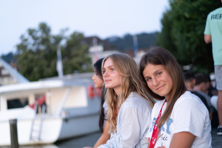 Two girls in white tops sit outside, one of them smiles straight to the camera, the other looks up to a direction.