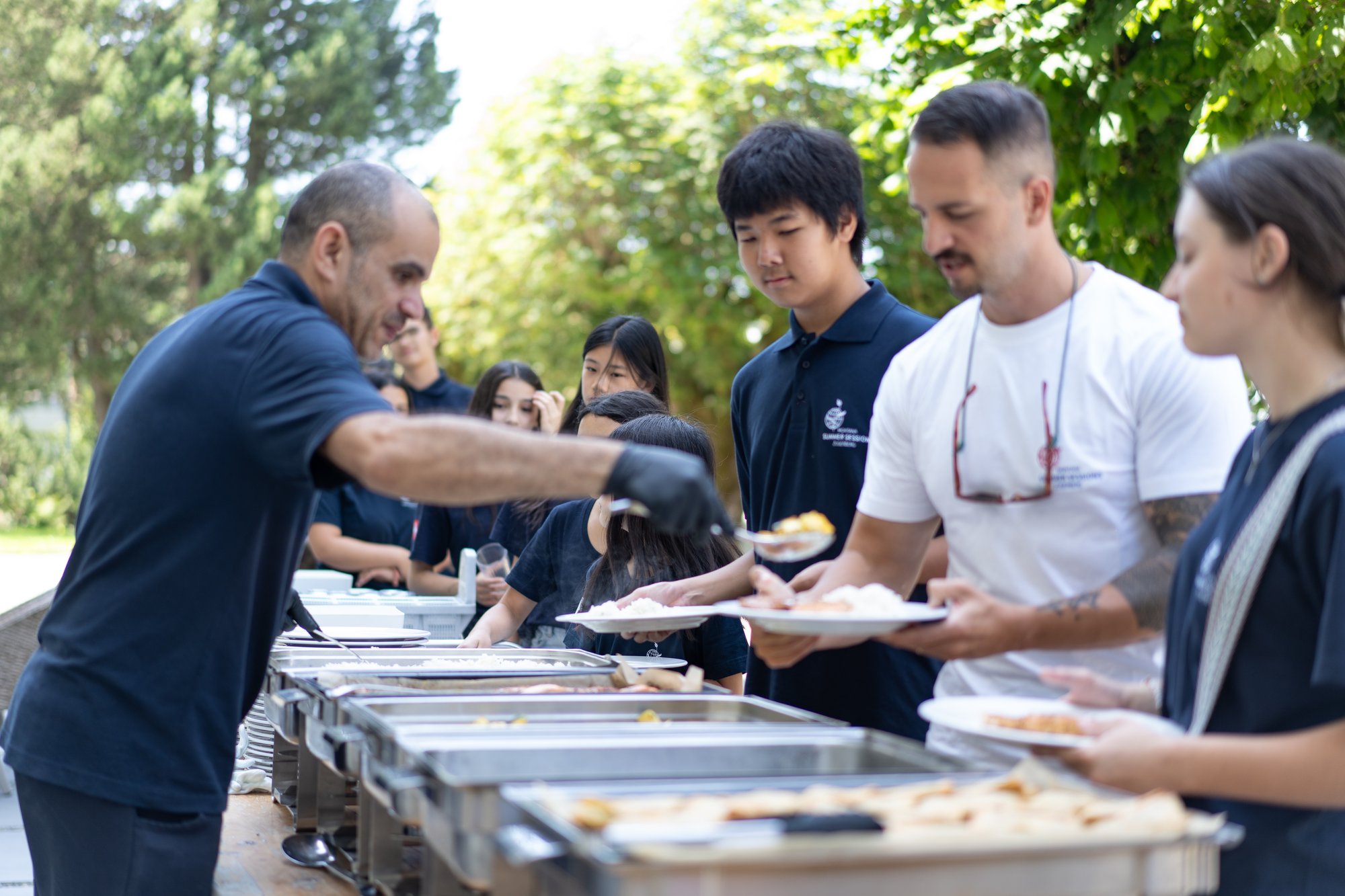 A male kitchen staff scoops some food on a plate of a man in an outdoor setting. There are other young kids nearby as well.