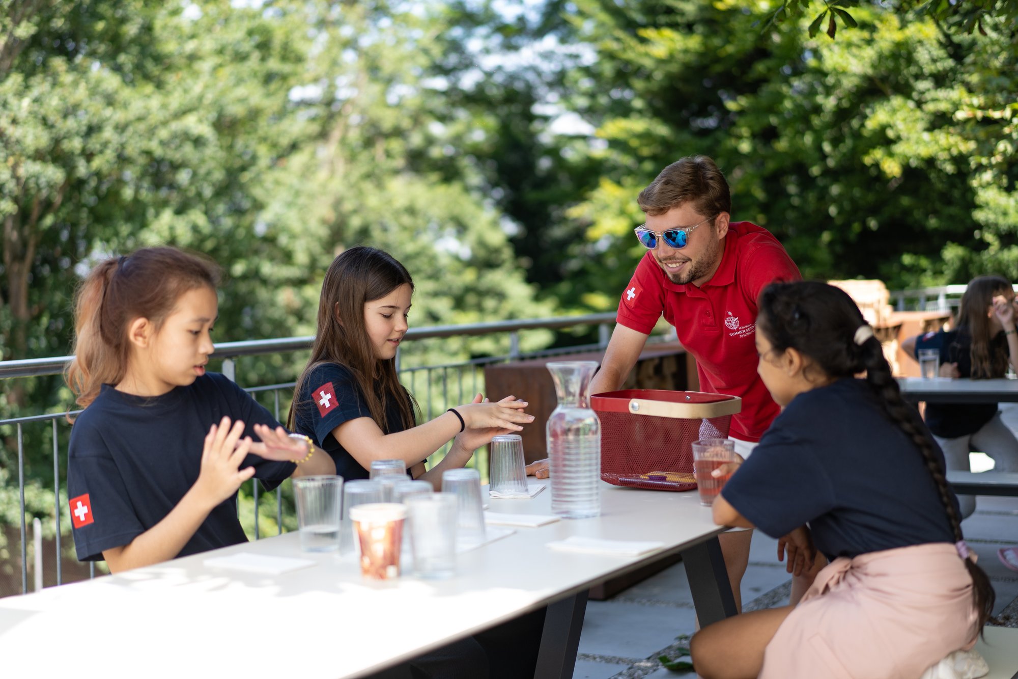 A young male mentor interacts with three girl students during an outdoor meal.