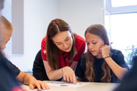 A female mentor helps a girl student with something, nearby there is a boy student as well.