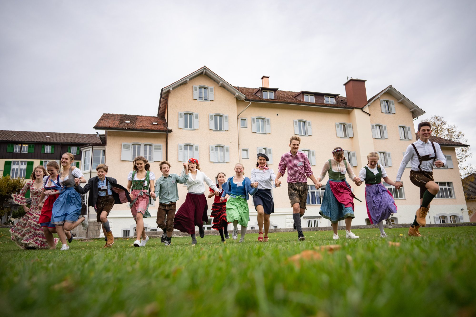 A group of boy and girl students in multicultural traditional outfits walk happily in front of a building.